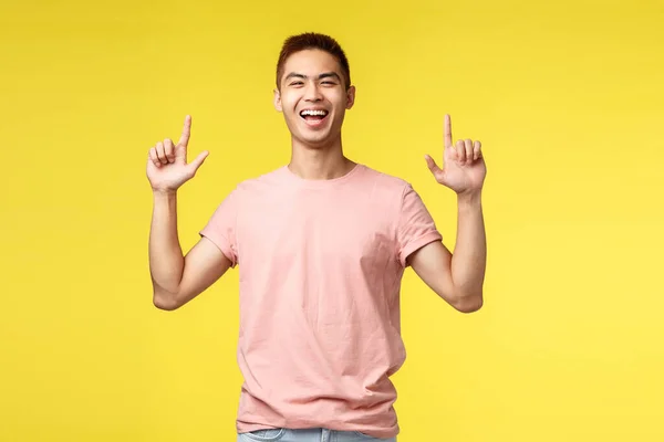 Mensen, verschillende emoties en levensstijl concept. Portret van een vrolijke knappe jongeman, Aziatische man die breed glimlacht, met de vingers omhoog wijst en lacht om de weg naar het product, nodigt uit tot een bezoek evenement — Stockfoto