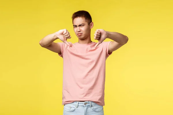 Horrível, muito decepcionante. Retrato de julgamento descontente asiático cara em rosa t-shirt, mostrar polegares para baixo e sorridente repugnante, sentir aversão, discordar, dar opinião negativa, fundo amarelo — Fotografia de Stock