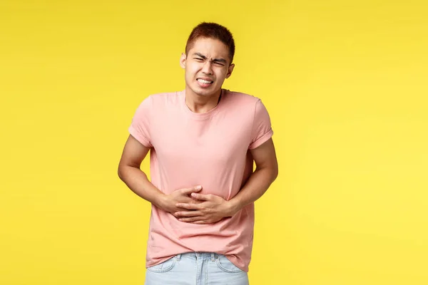 Saúde, pessoas e conceito de comida. Asiático cara em camiseta rosa, sentir-se doente, sorrindo de dor no estômago, de pé sobre fundo amarelo sofrem de envenenamento, de mãos dadas na barriga e flexão — Fotografia de Stock