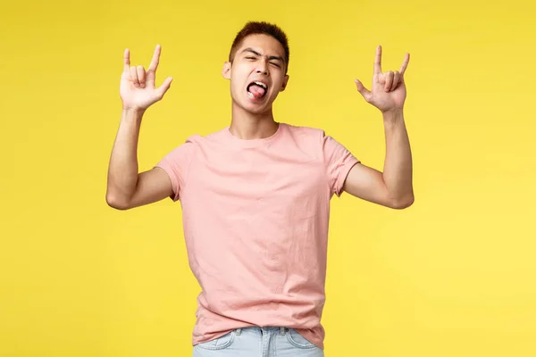 Estilo de vida, educação e conceito de pessoas. Entusiástico bonito e legal asiático homem em rosa t-shirt, mostrar língua e rock-n-roll sinal, se divertindo, festejando, de pé fundo amarelo — Fotografia de Stock