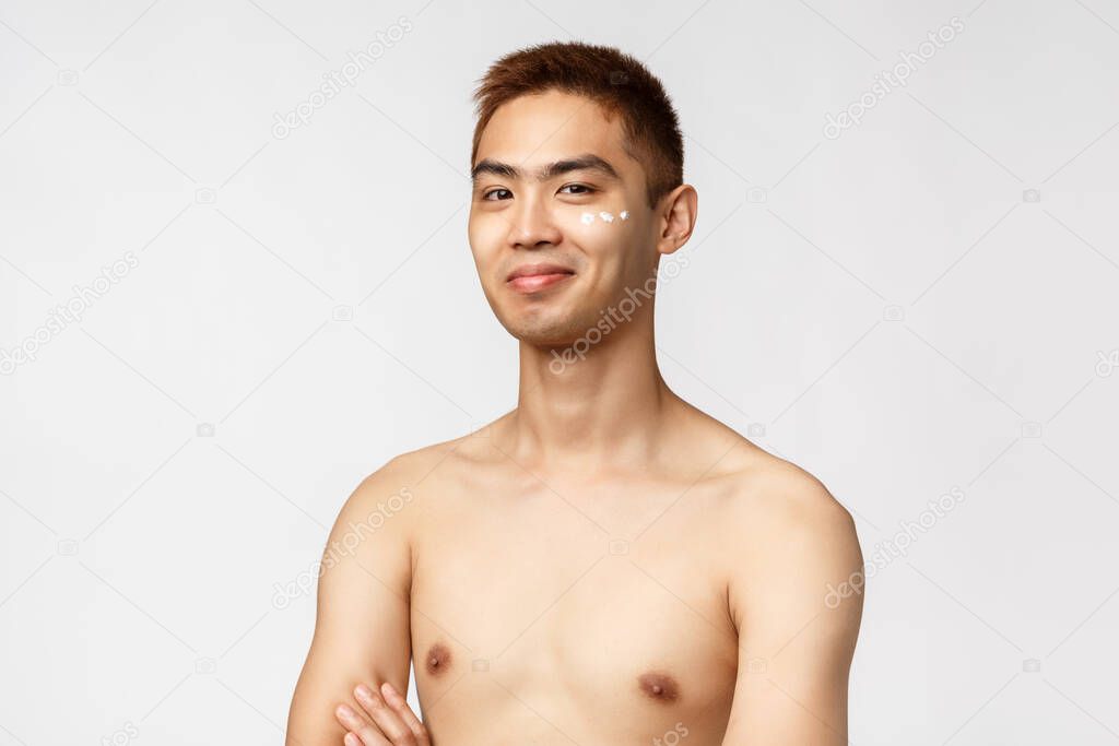 Beauty, people and leisure concept. Happy cheerful handsome asian man with naked torso look pleased, smiling apply skincare cosmetics, facial cream under eye, standing white background