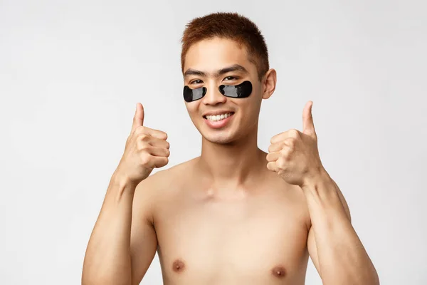 美，人和生活方式的概念。满意的亚洲裸体男子的画像，有眼斑，有大拇指向上的推荐标志，赞成和喜欢日常护肤产品，白色背景 — 图库照片