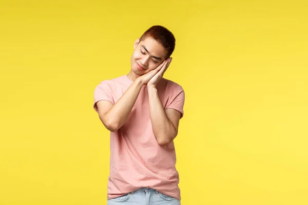 Lidé, různé emoce a životní styl. Zasněný asijský ospalý roztomilý kluk v růžovém tričku, snil o letních prázdninách, opírá se o dlaně jako usínající, usmívající se, žluté pozadí — Stock fotografie