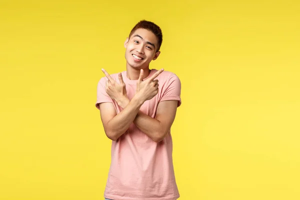 Hloupý a roztomilý asijský muž v růžovém tričku usmívající se mezi a starostlivý, nakloněná hlava půvabný jako ukazuje bokem na obě dobré varianty, ukazuje vlevo a vpravo, ukazuje dvě možnosti, stojí žluté pozadí — Stock fotografie