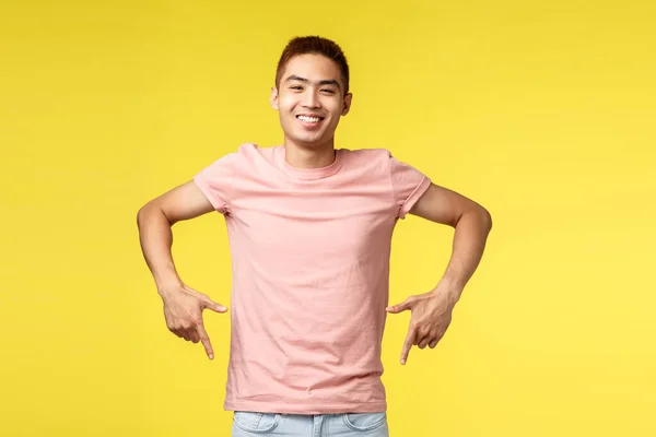 Retrato de easy-going alegre asiático cara em rosa t-shirt, convidando visita evento, apontando os dedos para baixo, sorrindo satisfeito, recomendo o produto, mostrando fundo promo, de pé fundo amarelo — Fotografia de Stock