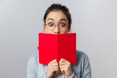 Eğitim, üniversite ve halk konsepti. Şaşırmış ve eğlendirilmiş Asyalı genç bir kızın yakın plan portresi, ilginç hikayede geniş gözler, kırmızı planlayıcının arkasındaki ağız, gri arka plan.