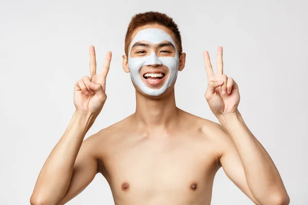 Szépség, bőr és spa koncepció. Vidám fiatal ázsiai srác meztelen törzs alkalmazott bőr rutin kozmetikumok, arc maszk, mutatják a béke jeleit, pihentető otthon, önellátó, fehér háttér — Stock Fotó