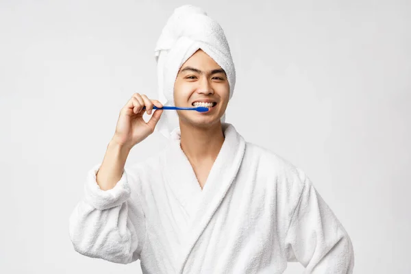 Szépség, spa és szabadidő koncepció. Vidám boldog ázsiai férfi fürdőköpenyben és törülközőben, fogmosás fogkefével és mosollyal, ügyelve a személyes higiénia, fehér háttér — Stock Fotó