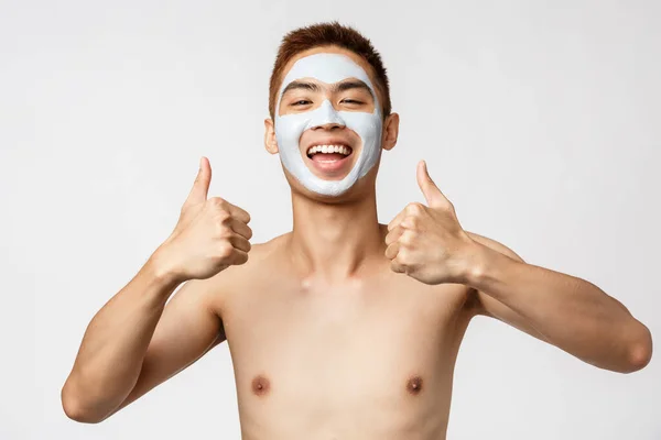 Szépség, bőr és spa koncepció. Boldog jóképű ázsiai férfi meztelen törzs arc maszk mosolygós és hüvelykujj-up, ajánljuk nagy bőr kozmetikai termék, fehér háttér — Stock Fotó