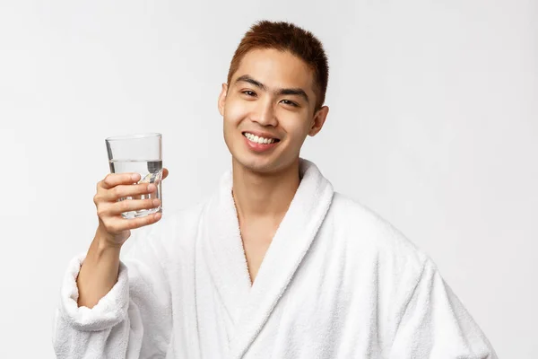 Szépség, spa és technológia koncepció. Igyunk az egészséges életmódra. Portré vidám ázsiai férfi fürdőköpeny emelése pohár víz és mosolygós, visel fürdőköpeny, ébredés és ital, fehér háttér — Stock Fotó