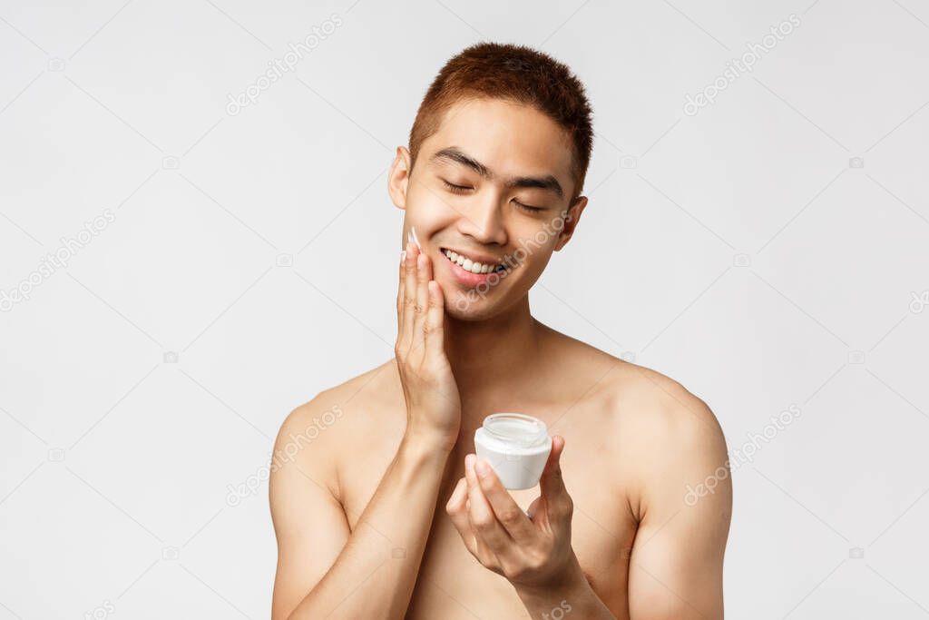 Beauty, people and leisure concept. Happy, silly handsome asian naked man close eyes, apply daily cream, smiling delighted as taking care of acne prone skin, standing white background