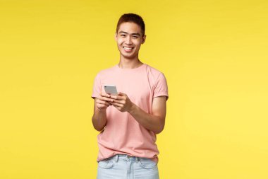 Teknoloji, iletişim ve yaşam tarzı konsepti. Neşeli, hevesli hippi adam portresi, pembe tişörtlü Asyalı erkek cep telefonu kullanıyor, kameraya gülüyor, çevrimiçi kalıyor, sipariş veriyor.