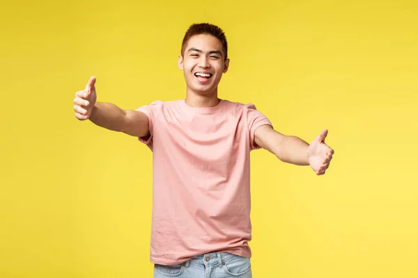 Lebensstil, Reise- und Personenkonzept. Porträt eines fröhlichen, freundlichen asiatischen Männchens, das zur Umarmung einlädt, die Hände nach Umarmungen ausstreckt, die Arme ausstreckt und breit lächelt, schmusend, vor gelbem Hintergrund stehend — Stockfoto