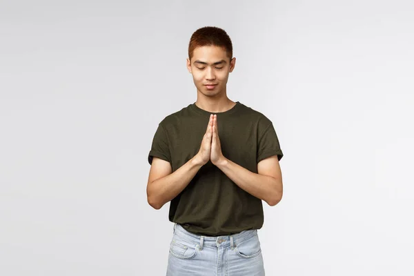 Retrato de esperançoso jovem asiático cara ter fé, fechar os olhos e sorrir, fechar as mãos juntos em oração, implorando a Deus, quer sonho tornado realidade, suplicando, de pé fundo cinza — Fotografia de Stock