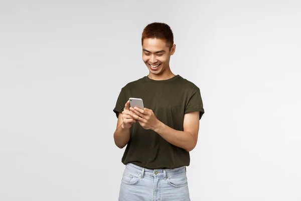 Teknoloji, online yaşam tarzı ve iletişim konsepti. Yeşil tişörtlü yakışıklı Asyalı bir gencin portresi. Neşeli bir şekilde cep telefonuna bakıyor, mesajlaşıyor, akıllı telefon ekranına gülümsüyor. — Stok fotoğraf