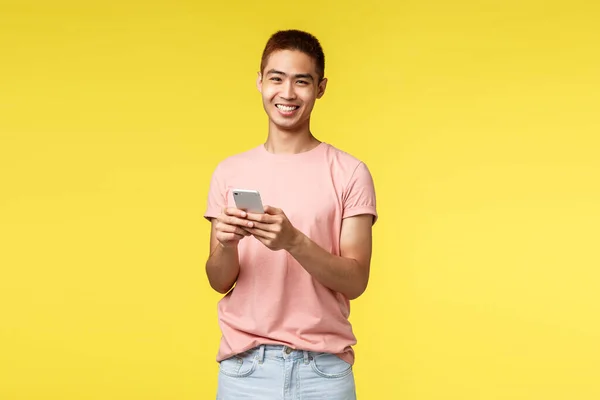 Технология, коммуникация и образ жизни. Портрет жизнерадостного, восторженного хипстера, азиатского мужчину в розовой футболке с помощью мобильного телефона, смеющегося над камерой, остающегося в сети, заказывающего доставку — стоковое фото