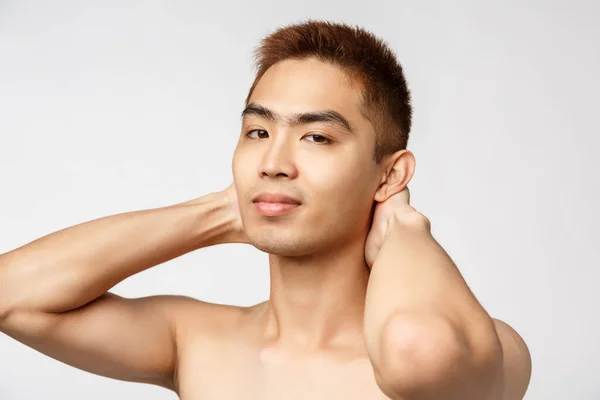 Beauty, skincare and men health concept. Portrait of handsome young asian man standing naked torso, hold hands behind head and smiling camera pleased, looking after appearance
