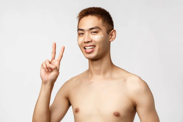 Krása, lidé a volnočasové pojetí. Fešák nadšený asijský muž s nahým trupem, ukázat mír znamení a usměvavý radost, aplikovat kosmetika krém pod okem a stát bílé pozadí — Stock fotografie