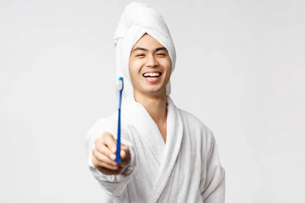 Szépség, spa és szabadidő koncepció. Fiatal boldog ázsiai férfi fürdőköpeny és törülköző, bemutató fogkefe és mosolygós vidám, ügyelve a személyes higiénia, álló fehér háttér — Stock Fotó
