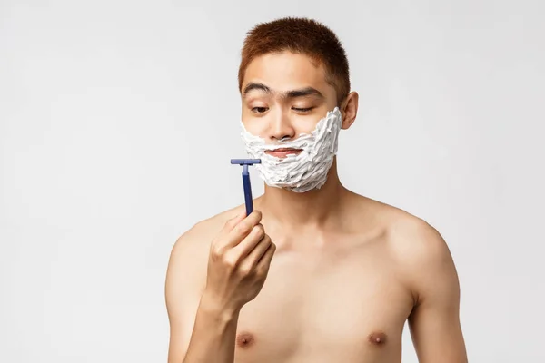 Szépség, emberek és higiénia. Kíváncsi jóképű ázsiai férfi néz új borotva, mint kezdő borotválkozás szakáll, álló krém az arcon és meztelen törzs, készülj fel a munkára, fürdőszoba rutin — Stock Fotó