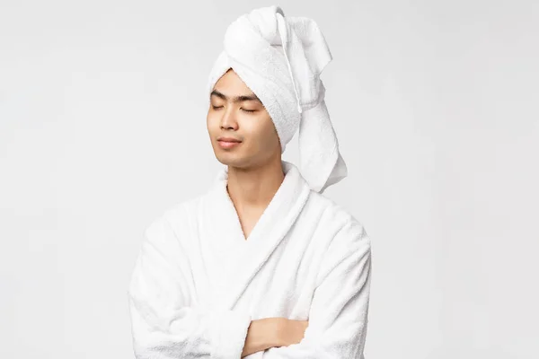 Piękno, spa i wypoczynek. Portret zrelaksowanego, spokojnego i spokojnego Azjaty cieszącego się wakacjami, zamkniętymi oczami i uśmiechniętym uśmiechem, noszącego szlafrok i ręcznik nad głową, białe tło — Zdjęcie stockowe