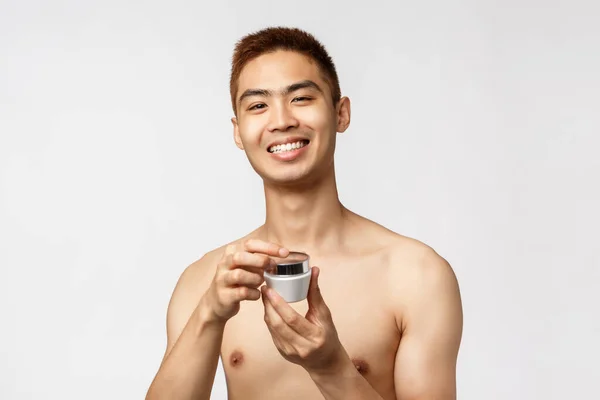 Belleza, gente y concepto de ocio. Guapo asiático desnudo hombre abierto crema para el cuidado de la piel, sonriendo complacido, aplicar el producto de rutina de la piel diaria y sonriendo satisfecho, teniendo cuidado de su propia cara, fondo blanco — Foto de Stock