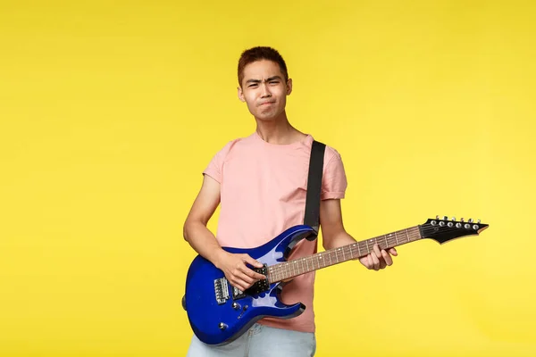 Lifestyle, leisure and youth concept. Enthusiastic carefree asian man in pink t-shirt playing on electric guitar over yellow background, enjoying performing with band, learn new instruments