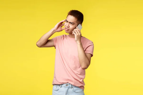 Tecnologia, comunicação e conceito de estilo de vida. perturbado asiático homem no rosa t-shirt, sinta-se frustrado, falando no celular, squinting e grimacing incomodado, ficar indeciso amarelo fundo — Fotografia de Stock