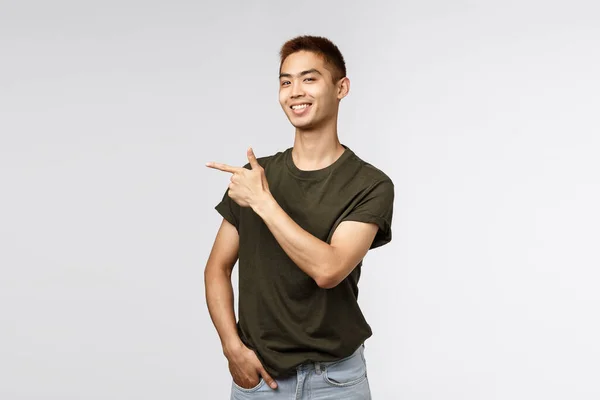 Orgulloso de buen aspecto asiático estudiante masculino señalando el dedo a la izquierda y sonriendo de la cámara complacido, chico mostrando el camino para almacenar, enlace en línea o bandera de la empresa, introducir el artículo, fondo gris —  Fotos de Stock