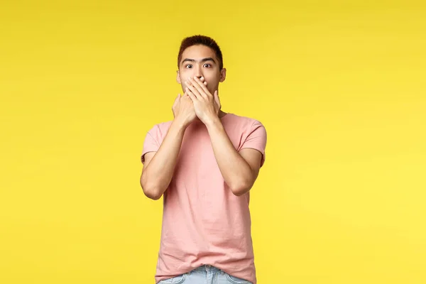 Люди, разные эмоции и образ жизни. Портрет шокированного азиатского хипстера в розовой футболке, задыхающегося от изумления, прикрывающего рот и выражающего изумление глазами, стоящего на жёлтом фоне — стоковое фото