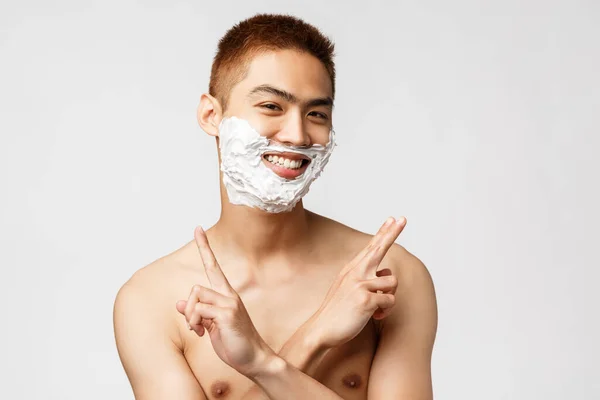 Szépség, emberek és higiénia. Portré vicces tinédzser ázsiai srác borotválkozás első alkalommal, alkalmazni krém szakáll és mosolygós, mutató béke jelek keresztbe kézzel, álló meztelen törzs — Stock Fotó