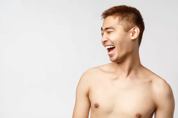 Belleza, gente y concepto de hogar. primer plano retrato de guapo joven asiático hombre con desnudo torso, mirando a la izquierda y riendo en voz alta, divertirse viendo hilarante rendimiento, fondo blanco — Foto de Stock