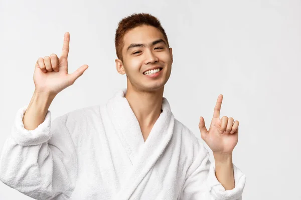Krása, lázně a volný čas koncept. Portrét veselého, pohledného nadšeného asijského muže v županu, ukazující prsty nahoru, ukazující cestu a tanec potěšen, radostně se usmívající, bílé pozadí — Stock fotografie