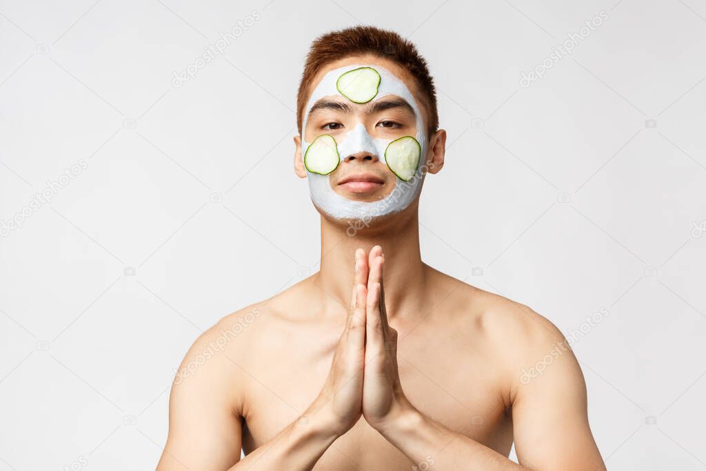 Beauty, skincare and spa concept. Portrait of relaxed, peaceful asian naked man with facial mask and cucumbers, make namaste gesture, clasp hands together over chest, white background
