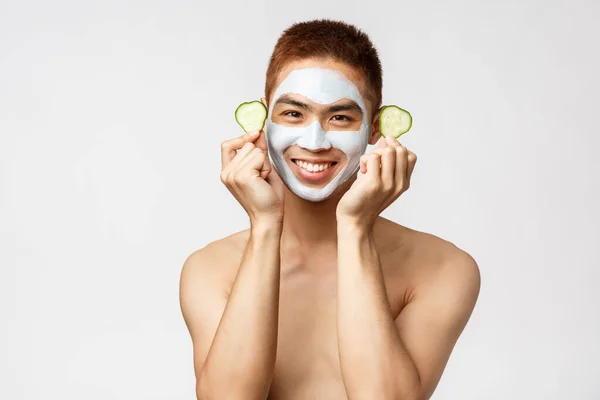 Schoonheid, huidverzorging en spa concept. Portret van knappe enthousiaste naakte aziatische man, met komkommer en glimlach in het gezicht masker, de behandeling van acne of puistjes, staan witte achtergrond — Stockfoto