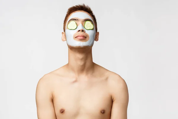 Bellezza, cura della pelle e concetto di spa. Ritratto di bell'uomo asiatico rilassato con busto nudo rilassante con maschera facciale e cetrioli sugli occhi, alzare la testa, sentirsi a proprio agio, in piedi sfondo bianco — Foto Stock