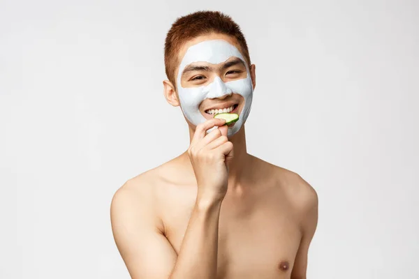 Szépség, bőr és spa koncepció. Jóképű ázsiai férfi meztelen törzs kozmetikai arc maszk, eszik uborka és mosolygós, pihentető szabadidő szalonban, álló fehér háttér — Stock Fotó
