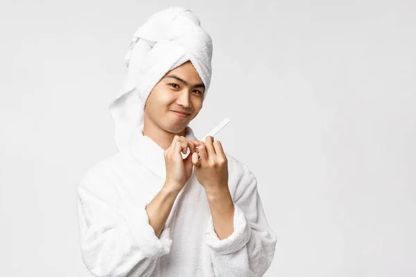 Концепция красоты, spa и отдыха. Портрет глупо красивый азиатский мужчина в халате и полотенце, улыбаясь приятно, полировка ногтей и сделать маникюр, стоя на белом фоне — стоковое фото