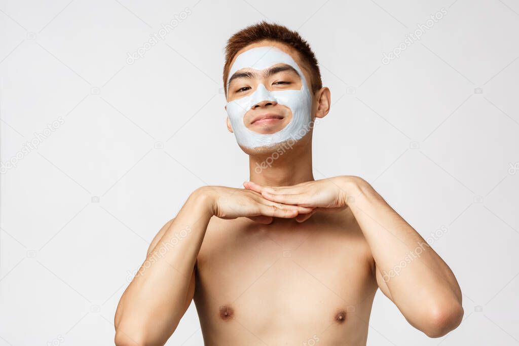 Beauty, skincare and spa concept. Portrait of lovely and silly naked asian man, smiling satisfied, wearing facial mask cream, cosmetic product taking care of skin, standing white background