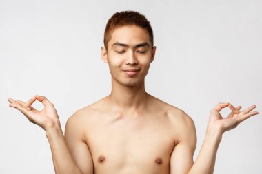 Güzellik, insanlar ve ev kavramı. Mutlu ve rahatlamış Asyalı bir adamın portresi. Beyaz arka planda çıplak duruyor, gözleri kapalı gülümsüyor, sakin ve sakin kalmak için meditasyon yapıyor.