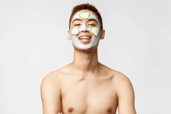 Szépség, bőr és spa koncepció. Közelkép portré boldog, nyugodt ázsiai férfi pihentető, álló fürdőszoba meztelen törzs mosolygó, alkalmazni arc maszk és uborka az arcon, fehér háttér — Stock Fotó
