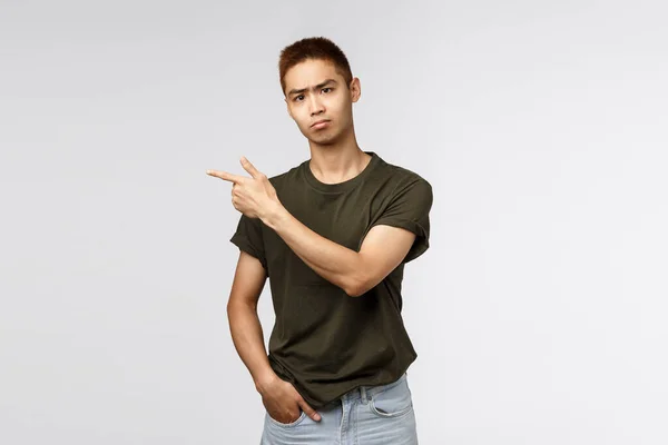 Portrét skeptického, zachmuřeného mladého asijského studenta nesnáší sebedistancování se od přátel a spolužáků z vysoké školy, ukazování prstem vlevo a šklebení fotoaparátu, trucování rozčilením, závist nebo lítost — Stock fotografie
