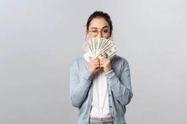 Business, finance and investment concept. Happy, pleased pretty asian woman, rich girl playfully raise eyebrow hiding satisfied smile behind money dollars, win cash prize, grey background — Stock Photo, Image