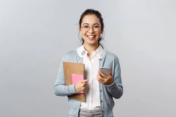 Oktatás, tanárok, egyetemi és iskolai koncepció. Vidám okos fiatal női oktató, gazdaság okostelefon és notebook könyvekkel, előkészítése leckék, tananyag, szürke háttér — Stock Fotó