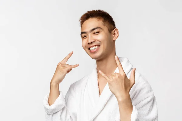Beauty, Wellness- und Freizeitkonzept. Porträt eines coolen, gut aussehenden asiatischen Mannes im Bademantel, mit Rock-n-Roll-Zeichen, Schwermetall, zufrieden lächelnd, Zeit an heißen Quellen genießen, Urlaubszeit — Stockfoto