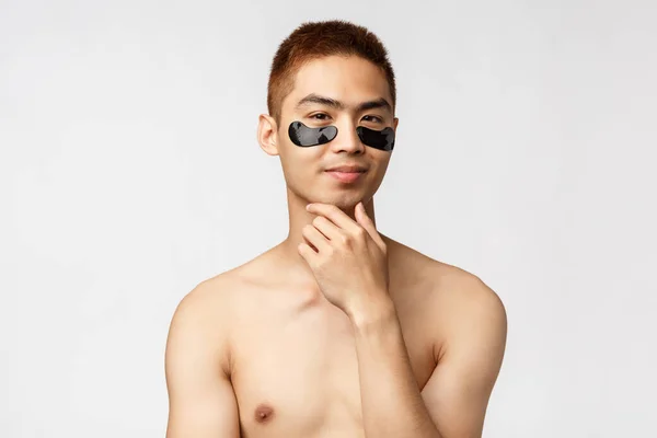 Beauty, people and lifestyle concept. Thoughtful enthusiastic, handsome naked asian man wearing eye-patches and smiling, have an idea, thinking, consider apply skincare mask, white background — Stock Photo, Image