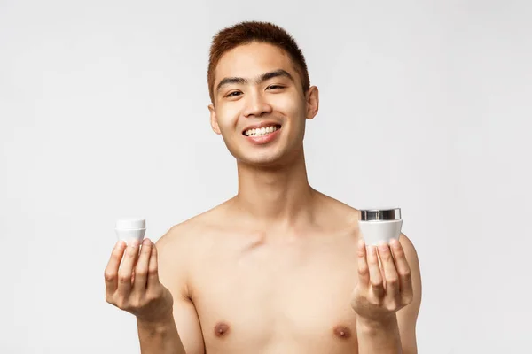 Beauty, people and leisure concept. Portrait of enthusiastic, handsome man in naked torso, holding two bottles of facial cream and smiling pleased, taking care of skin blemishes, white background