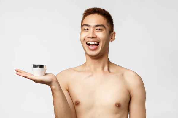 Belleza, gente y concepto de ocio. Retrato de alegre sonriente hombre asiático con el torso desnudo introducir su rutina favorita cuidado de la piel, anti-manchas crema, sosteniendo tarro en la palma y mirada entusiasta — Foto de Stock