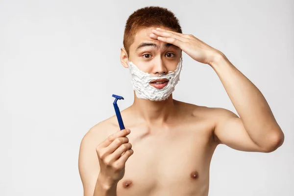 Szépség, emberek és higiénia. Összezavarodott aranyos ázsiai tini srác próbál borotválkozni első alkalommal, úgy érzi, egy kicsit ideges, megható homlok, mutatja borotva és alkalmazni borotvahab, fehér háttér — Stock Fotó