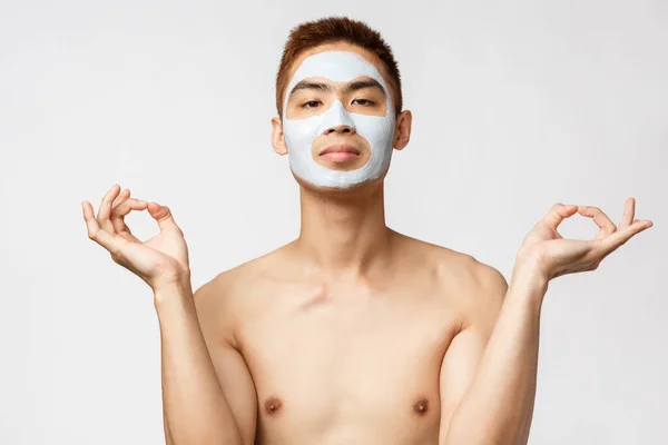 Szépség, bőr és spa koncepció. Portré nyugodt fiatal ázsiai férfi arc anti-akne maszk, kozmetikai termék, meditál, hogy zen jel do jóga, úgy néz ki, békés, élvezi otthon maradni — Stock Fotó